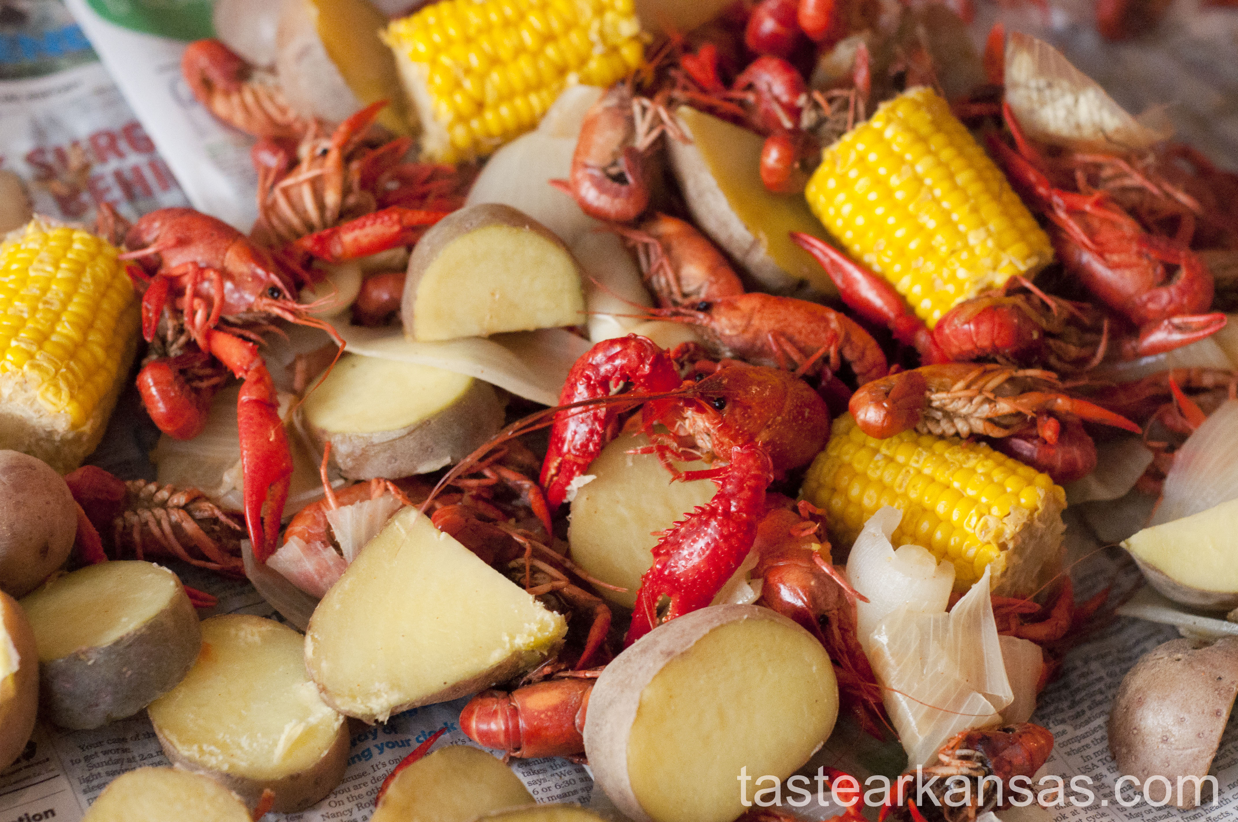 this image is of a crawfish boil, with crawfish, corn, onions, potatoes and garlic on a newspaper covered table